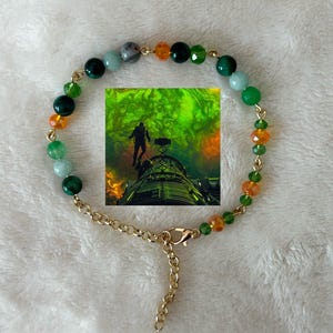 May include: A beaded bracelet with a gold-colored chain and clasp. The bracelet features a variety of colorful beads, including green, orange, and gray. A small, framed image is centered on the bracelet.