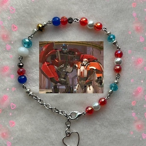 Peut inclure: Un bracelet de perles avec une chaîne argentée et un charm en forme de cœur. Le bracelet présente des perles rouges, bleues, blanches et irisées. Une petite image de deux robots est au centre.