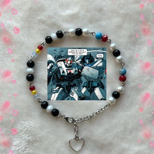 May include: A beaded bracelet featuring black, white, red, yellow, and blue beads. A small comic strip is centered on the bracelet, with the text "WHY IS IT HE'S AFRAID OF YOU BUT NOT OF ME? TONE." A silver heart charm dangles from the clasp.