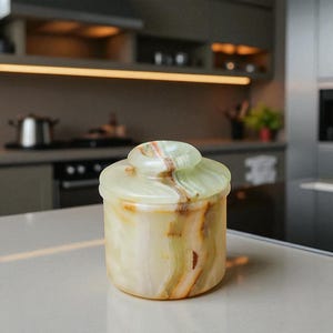 May include: A small, cylindrical jar with a lid, crafted from onyx. The jar displays a natural, marbled pattern in shades of light green and brown. The lid mirrors the design. The container is placed on a white surface.