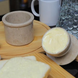 May include: A beige marble butter dish with a matching lid, filled with yellow butter, sits on a wooden board. A white mug and a decorative glass are in the background. Slices of bread are in the foreground.