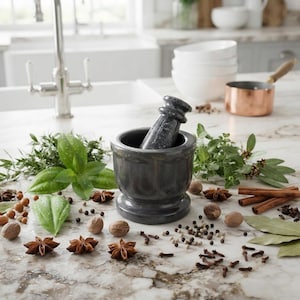 Può includere: Un mortaio con pestello in pietra grigio scuro circondato da spezie intere sparse tra cui anice stellato, noce moscata, grani di pepe, chiodi di garofano, stecche di cannella e foglie di alloro, insieme a erbe verdi fresche su marmo.