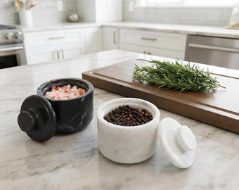 May include: Two cylindrical stone salt and pepper cellars with matching lids. One cellar is black marble filled with pink Himalayan salt, and the other is white marble filled with whole black peppercorns. Both have circular knob handles.