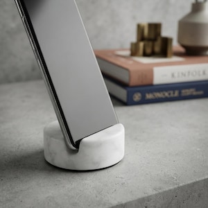 May include: A circular white marble phone stand with grey veining, designed to hold a smartphone upright. The stand features a smooth, polished finish and a recessed slot for the device, providing a stable base for a mobile phone on a flat surface.