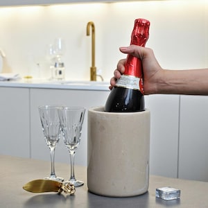Può includere: Un refrigeratore per vino cilindrico in pietra beige, due flûte da champagne in vetro trasparente con linee verticali incise, un apribottiglie in metallo dorato e un cubetto di ghiaccio quadrato. Una bottiglia di spumante con capsula rossa è inserita.