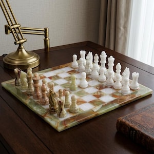 May include: A chess set with a green, white, and brown marble chessboard. The chess pieces are white and brown. The board is set up and ready to play. A gold-colored lamp is in the background.