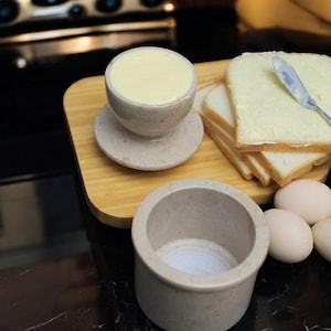 May include: A stone butter dish filled with yellow butter, on a wooden board beside buttered bread and a knife. A small stone bowl with salt and several eggs are also present.