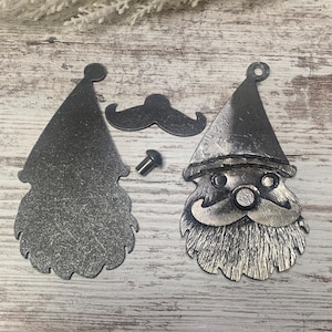 May include: Metal Santa Claus face and hat cutouts with a separate mustache and a small screw. The Santa face has a textured beard and hat. The metal has a dark, weathered appearance, suggesting an aged or rustic aesthetic. Ideal for holiday crafts.