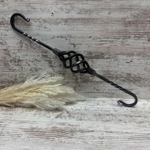 May include: A black wrought iron plant hanger with a decorative spiral design in the center and curved hooks on each end. The metal has a twisted design on the stem. The background is a light wood.
