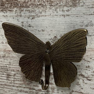 May include: A bronze butterfly wall hook with a textured finish. The hook is in the shape of a butterfly with its wings spread out. The hook is attached to a white wooden surface.