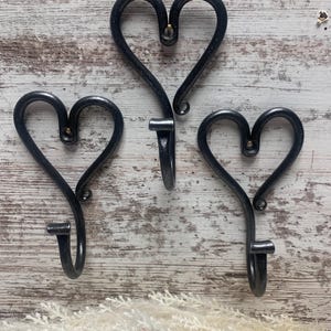 May include: Three wrought iron heart-shaped wall hooks. The hooks are dark gray with a matte finish and are mounted on a light-colored wooden surface. Each hook has a heart shape at the top and a curved hook at the bottom.