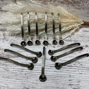 May include: An assortment of antique-style metal spoons with small, round bowls and long, slender handles. The spoons have a dark, aged finish, and are arranged on a light-colored wooden surface.