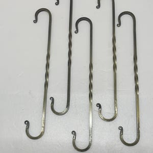 May include: Set of five handcrafted metal hooks. Each hook has a curved top and bottom, with a twisted design on the stem. The hooks are a dark, metallic color, and are likely made of iron or steel.