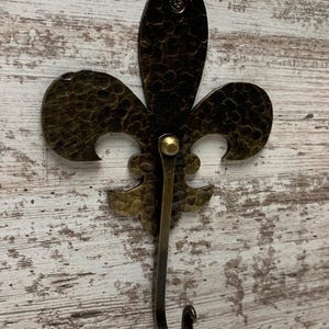 May include: A hammered brass fleur-de-lis hook with a decorative design. The hook is mounted on a white wooden surface.