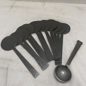 May include: A collection of dark gray metal spoons with round bowls and long, rectangular handles. One spoon has a larger, rounded bowl. The spoons are arranged on a white surface, suggesting a kitchen or dining theme.