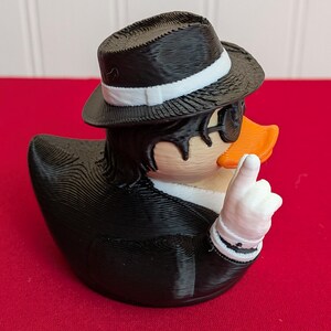 May include: A novelty rubber ducky figurine dressed in a black suit, fedora, and sunglasses. The duck has an orange beak and is giving a thumbs-up gesture with a white-gloved hand. The duck is on a red surface.