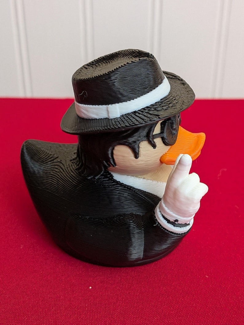 May include: A novelty rubber ducky figurine dressed in a black suit, fedora, and sunglasses. The duck has an orange beak and is giving a thumbs-up gesture with a white-gloved hand. The duck is on a red surface.