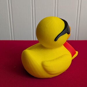 May include: A yellow rubber duck with black sunglasses and a red scarf. The duck is sitting on a red surface, with a white background. The duck is a novelty item.