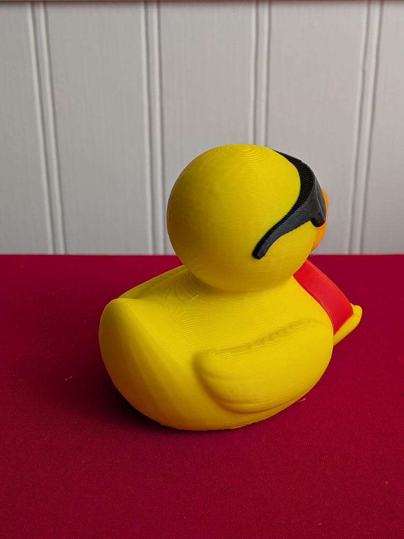 May include: A yellow rubber duck with black sunglasses and a red scarf. The duck is sitting on a red surface, with a white background. The duck is a novelty item.