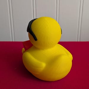 May include: A yellow rubber duck toy with a black headband and a red object. The duck is sitting on a red surface, with a white wall in the background. The duck is a novelty item.
