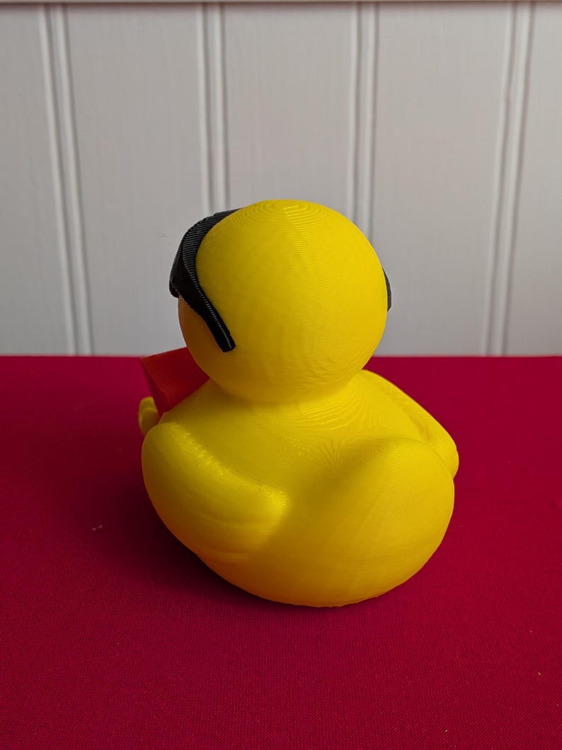 May include: A yellow rubber duck toy with a black headband and a red object. The duck is sitting on a red surface, with a white wall in the background. The duck is a novelty item.