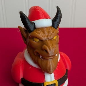 May include: A 3D-printed figurine of a devil in a Santa Claus outfit. The figure has a brown face, yellow eyes, black horns, and is wearing a red Santa suit with a black belt and gold buckle. The Santa hat is red with a white trim.