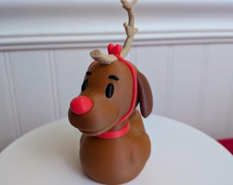 May include: A brown reindeer figurine with a red nose, collar, and headband. The reindeer has black eyes and small antlers. The figurine is sitting on a white surface.