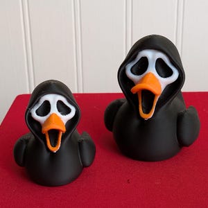 May include: Two rubber duck figures in different sizes, each featuring a black hooded cloak, a white mask with hollow eye sockets, and an orange beak. The ducks have a matte finish and are positioned side by side, displaying a consistent design.