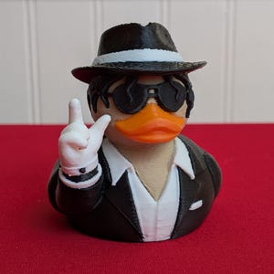 May include: A 3D-printed rubber ducky wearing a black fedora with a white band, sunglasses, and a black suit with a white shirt. The duck is making a hand gesture with a white-gloved hand. The duck has an orange beak.