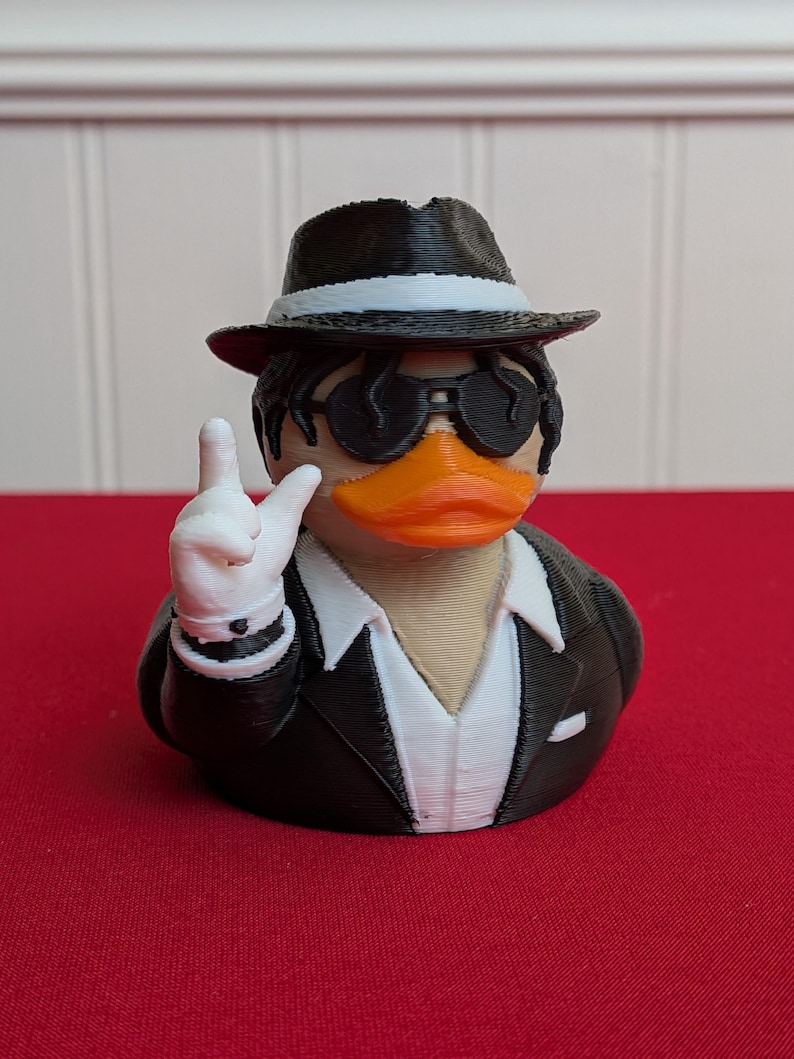 May include: A 3D-printed rubber ducky wearing a black fedora with a white band, sunglasses, and a black suit with a white shirt. The duck is making a hand gesture with a white-gloved hand. The duck has an orange beak.
