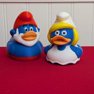 May include: Two novelty duck figurines, one male and one female, in the style of Smurfs characters. The male duck has a red hat and white beard, while the female duck has a white hat and yellow hair. Both ducks are blue with orange beaks.