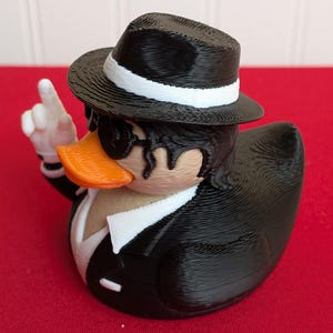 May include: A novelty rubber ducky dressed in a black suit, fedora with a white band, and sunglasses. The duck has an orange beak and is giving a hand gesture. The background is red.