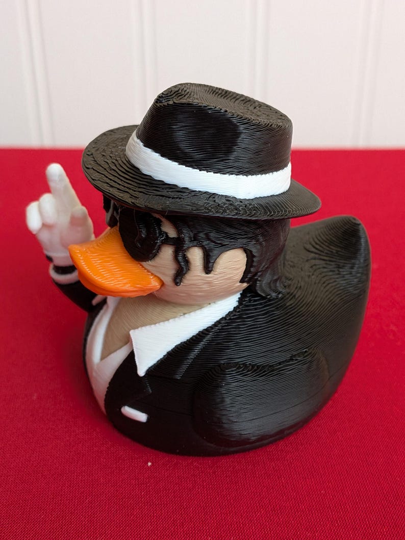 May include: A novelty rubber ducky dressed in a black suit, fedora with a white band, and sunglasses. The duck has an orange beak and is giving a hand gesture. The background is red.