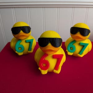 May include: Three yellow rubber ducks wearing black sunglasses and the number 67. The number 67 is in green on two ducks and red on the other. The ducks are on a red surface.