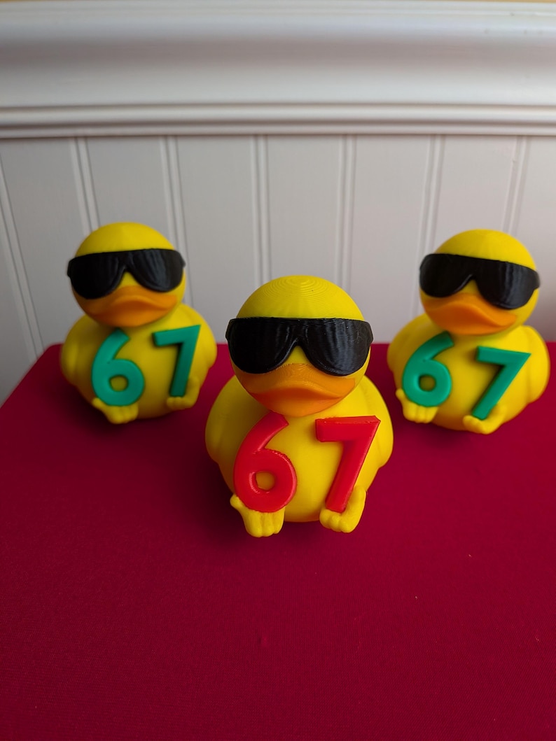 May include: Three yellow rubber ducks wearing black sunglasses and the number 67. The number 67 is in green on two ducks and red on the other. The ducks are on a red surface.