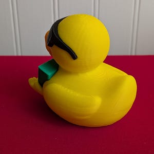 May include: A bright yellow duck figurine wearing black sunglasses and holding a small green block. The duck has an orange beak and is sitting on a red surface. The figurine is a whimsical decorative item.