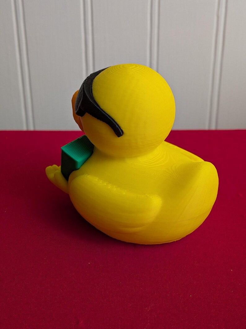 May include: A bright yellow duck figurine wearing black sunglasses and holding a small green block. The duck has an orange beak and is sitting on a red surface. The figurine is a whimsical decorative item.