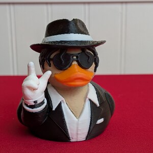 May include: A 3D-printed duck figurine wearing a black fedora, sunglasses, and a black suit with a white shirt. The duck is making a peace sign with its white-gloved hand. The duck has an orange beak and is set against a red background.