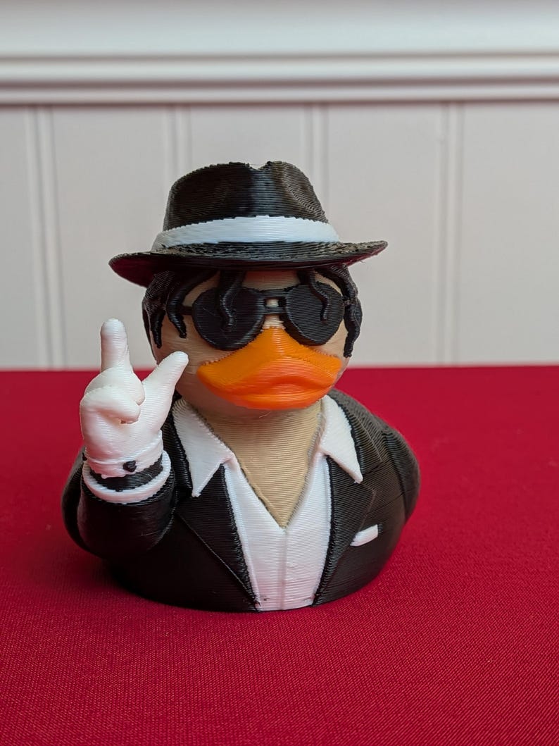 May include: A 3D-printed duck figurine wearing a black fedora, sunglasses, and a black suit with a white shirt. The duck is making a peace sign with its white-gloved hand. The duck has an orange beak and is set against a red background.