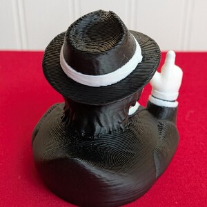 May include: A black and white figurine of a duck wearing a fedora with a white band and a black suit. The duck is giving the middle finger with a white-gloved hand. The figurine is on a red surface.