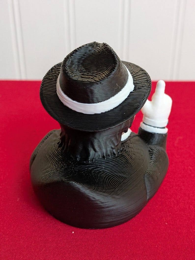 May include: A black and white figurine of a duck wearing a fedora with a white band and a black suit. The duck is giving the middle finger with a white-gloved hand. The figurine is on a red surface.