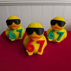 May include: Three yellow rubber ducks wearing black sunglasses. Each duck has the number "67" in either red or green. The ducks are on a red surface.