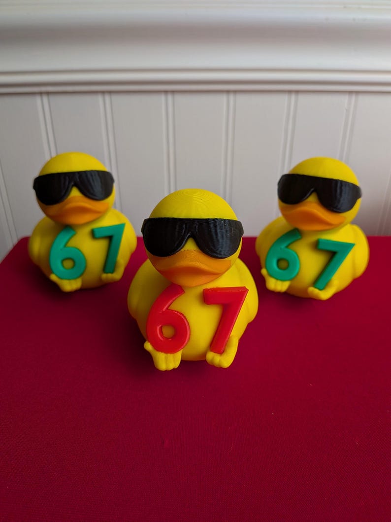 May include: Three yellow rubber ducks wearing black sunglasses. Each duck has the number "67" in either red or green. The ducks are on a red surface.