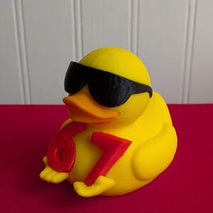 May include: A yellow rubber duck wearing black sunglasses and holding the numbers 6 and 7 in red. The duck has an orange beak and is sitting on a red surface.