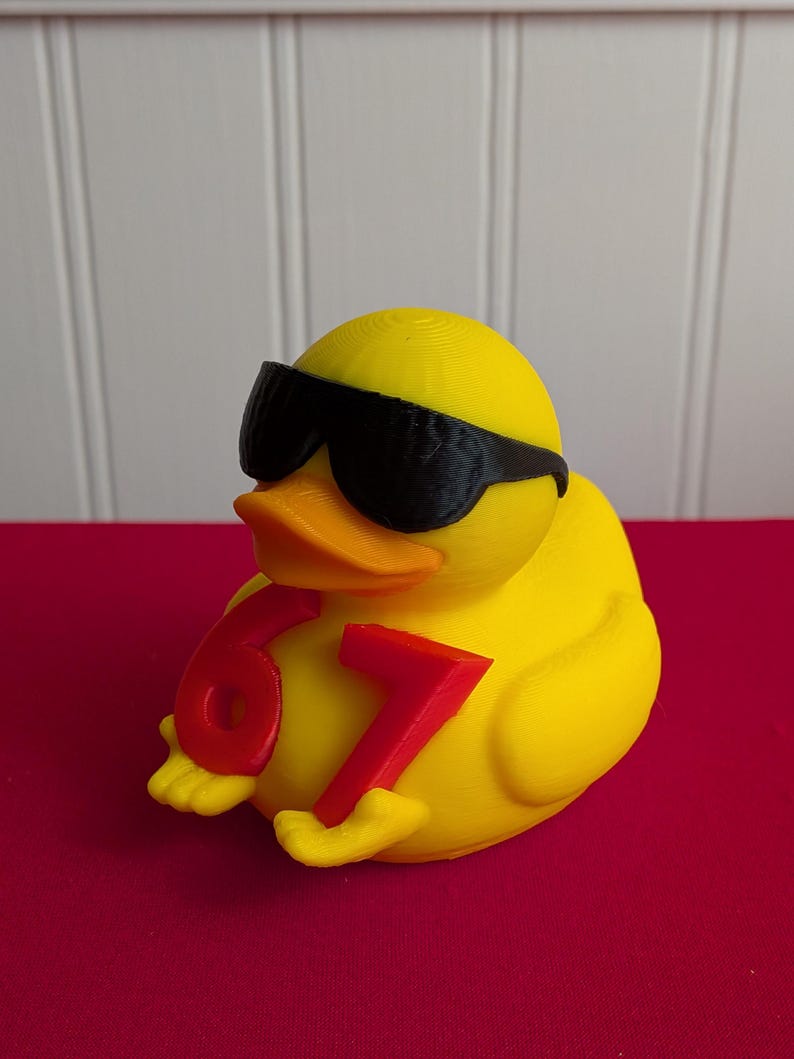 May include: A yellow rubber duck wearing black sunglasses and holding the numbers 6 and 7 in red. The duck has an orange beak and is sitting on a red surface.