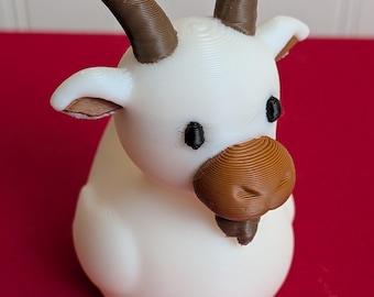 May include: A 3D printed white goat figurine featuring brown horns, a brown snout, a small brown beard, and black eyes. The surface of the object displays visible horizontal layer lines from the printing process, giving it a textured appearance.