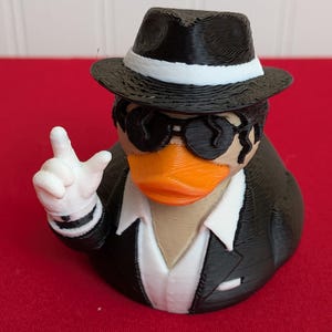 May include: A novelty rubber ducky figurine dressed in a black suit, fedora, and sunglasses. The duck has an orange beak and is giving a hand gesture with a white-gloved hand. The duck is on a red surface.