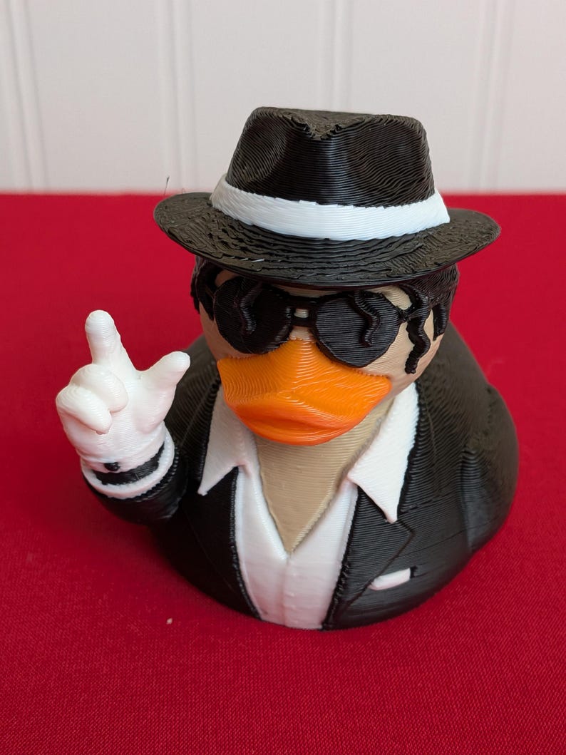 May include: A novelty rubber ducky figurine dressed in a black suit, fedora, and sunglasses. The duck has an orange beak and is giving a hand gesture with a white-gloved hand. The duck is on a red surface.