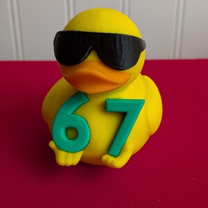 May include: A yellow rubber duck wearing black sunglasses and holding the number 67 in green. The duck has an orange beak and is set against a red background.