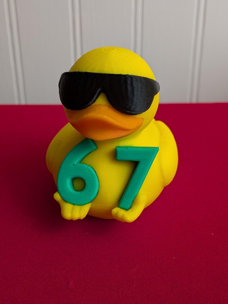 May include: A yellow rubber duck wearing black sunglasses and holding the number 67 in green. The duck has an orange beak and is set against a red background.